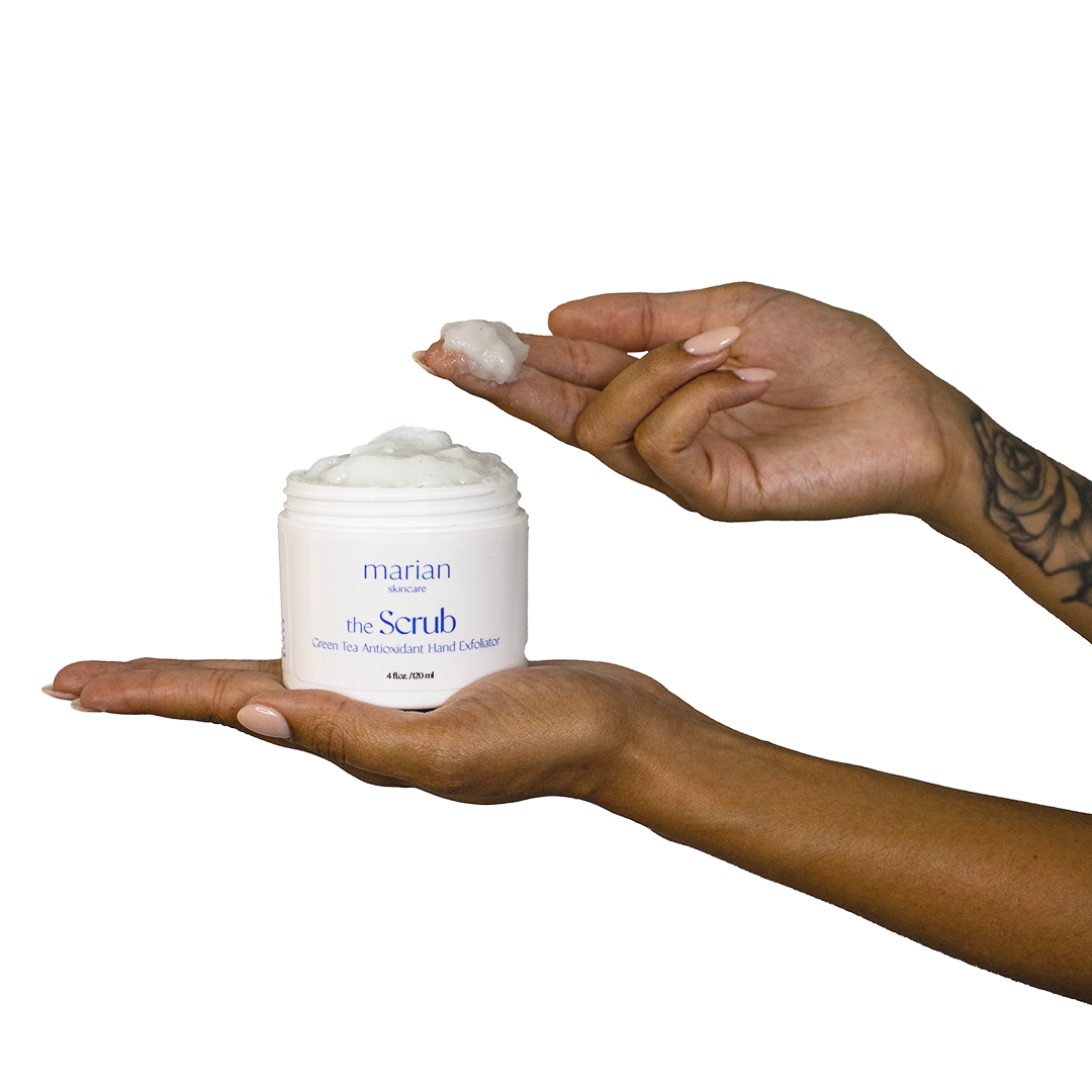 A model holds a jar of marian skincare the Scrub Green Tea Antioxidant Hand Exfoliator in the open palm of their left hand. The model has scooped a small amount of the scrub onto their right index and middle fingers.