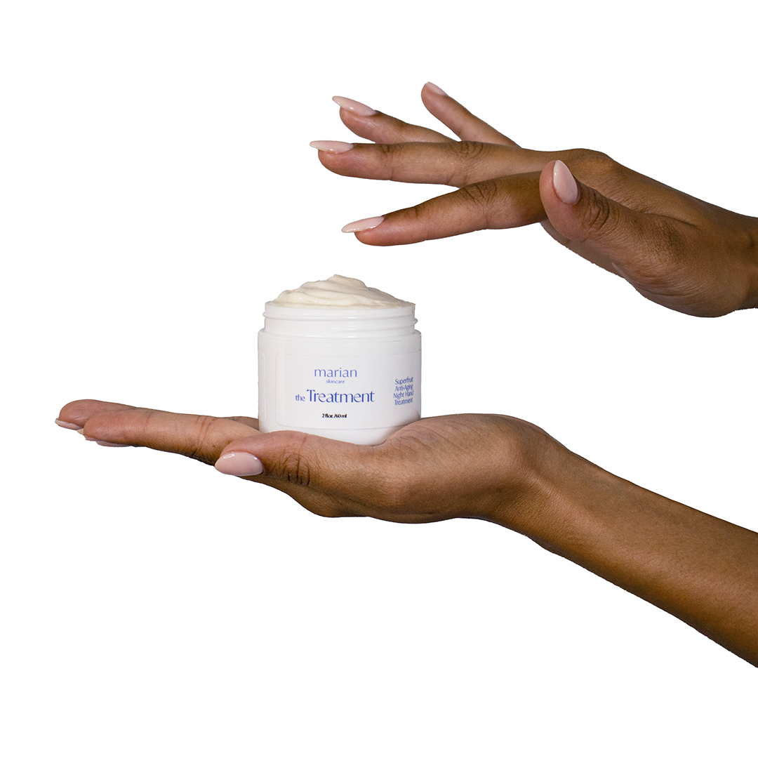A model holds a jar of marian skincare the Treatment Superfruit Anti-Aging Night Hand Treatment in the palm of their left hand. Their right hand hovers over the open jar with their index finger reaching down to graze the product in the jar.